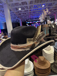 Black felt western hat with beige trim, frayed fabric band, decorative feathers and metal concho on display at an indoor market or event with stacks of cowboy hats and colorful stage lights in the background.