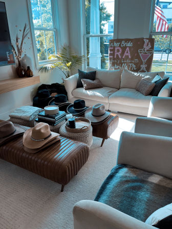 Sunlit Nashville living room with cream sectional, leather ottomans and a collection of wide-brim hats on coffee tables, potted palm, decorative pillows and tall windows showing an American flag outside.