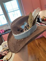 Brown felt cowboy hat with light-blue frayed band, gold star charm trim, decorative feathers and vintage card tucked in, held over a pink kitchen with hardwood floor