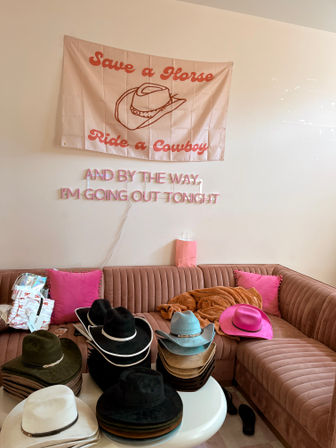 Stacks of colorful cowboy hats on a white table and pink velvet sectional sofa under a 'Save a Horse Ride a Cowboy' wall flag and pink neon sign in a pastel boutique-style room.