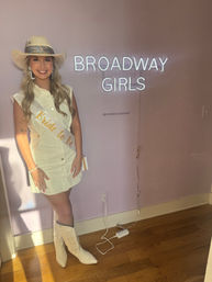 Smiling bride-to-be in a cowboy hat, white sash and sleeveless dress with white embroidered cowboy boots posing indoors against a pink wall with a neon "BROADWAY GIRLS" sign
