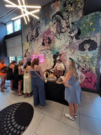 People browsing a cowboy hat pop-up inside a venue in front of a colorful Nashville-inspired mural of cowgirls, horses and neon accents