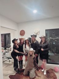 Seven women in cowboy hats and boots pose for a cowgirl-themed party in a modern city apartment with wall-mounted hats, cowhide rug, pink balloons and a nighttime skyline view.