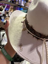 White felt cowboy hat with feather trim and studded leather band featuring small gold cactus and letter B charms, held by a hand wearing a green "PARTNER" wristband at a craft fair vendor booth.