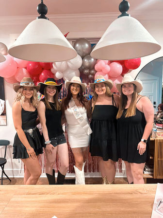 Five women in cowgirl hats and boots posing indoors for a festive party with pink and silver balloon garland and pendant lights, the center guest wearing a white dress and sash.