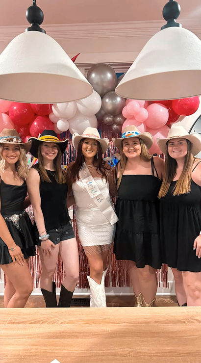 Five women in cowgirl hats and boots posing indoors for a festive party with pink and silver balloon garland and pendant lights, the center guest wearing a white dress and sash.