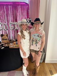 Two smiling women in cowgirl outfits — cowboy hats and boots — at an indoor pink-fringe backdrop party or pop-up; one wears a white button dress with white hat and boots, the other sports a graphic tee, leopard shorts, black hat and brown boots, standing beside a vendor table with accessories and metallic letter balloons.