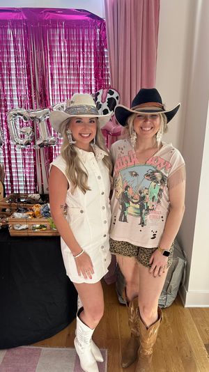 Two smiling women in cowgirl outfits — cowboy hats and boots — at an indoor pink-fringe backdrop party or pop-up; one wears a white button dress with white hat and boots, the other sports a graphic tee, leopard shorts, black hat and brown boots, standing beside a vendor table with accessories and metallic letter balloons.