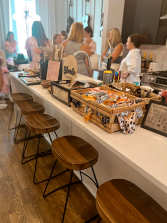 Modern kitchen island with wooden bar stools, crates of colorful hair scarves and accessories on the countertop, and a group of people chatting in the background — cozy pop-up gathering vibe.