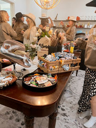 Indoor boho‑western hat bar with guests trying on cowboy hats around a table covered in hats, feathers, bandanas, floral arrangements and trays of hat pins and accessories.