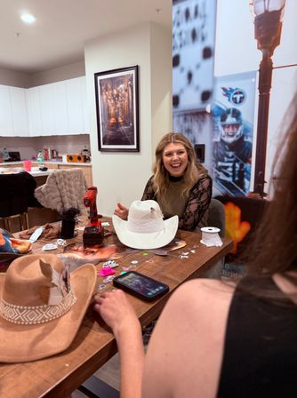 Smiling woman at a wooden table during a cowboy-hat craft night, decorating white and tan hats with ribbons, feathers, sequins, a drill and phone in a modern apartment kitchen.