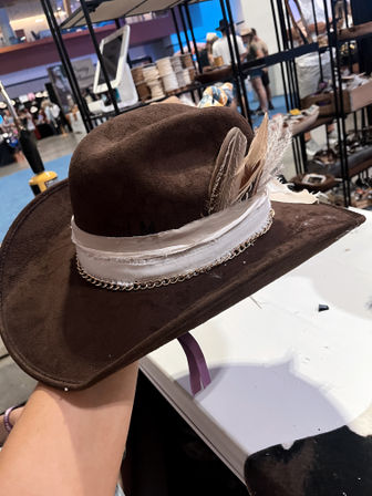 Brown suede cowboy hat with a white fabric band, chain trim and feather accents displayed on a table at an indoor artisan market booth