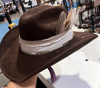 Brown suede cowboy hat with a white fabric band, chain trim and feather accents displayed on a table at an indoor artisan market booth