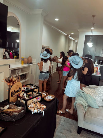 Group of women trying on cowboy-style and wide-brim hats at a home pop-up hat bar in a modern living room and kitchen with hat racks and accessory table