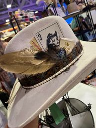 White felt cowboy hat with gold feather and feathered band, Queen of Spades card and "We do it right" tag, displayed at a craft market booth.
