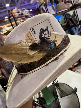 White felt cowboy hat with gold feather and feathered band, Queen of Spades card and "We do it right" tag, displayed at a craft market booth.