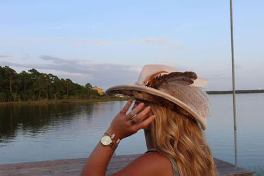 Person in a feathered cowboy hat and gold watch on a wooden dock, gazing across a calm lake with tree-lined shore under a pastel evening sky.