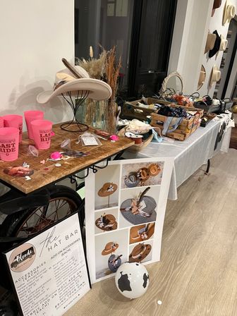 Indoor boho hat bar for a bridal party — cowboy and felt hats on stands, wooden crates of ribbons, feathers and trims, dried floral vase, pink 'Bride’s Last Ride' cups, styling poster and accessories on a wooden table.