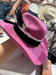 Bright pink felt cowboy hat with a black feather, striped band and gold chain trim on display at a busy market table
