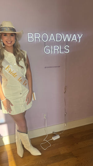 Smiling woman in a white cowboy hat, cream button-front dress, "Bride to Be" sash and white cowboy boots posing against a pink wall with a glowing neon sign and hardwood floor.