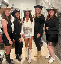 Five women in cowgirl-chic outfits wearing cowboy hats and boots, smiling and posing indoors in a modern hallway with patterned wallpaper and wood floor; center woman in a white blazer, silver mini skirt and white cowboy boots.