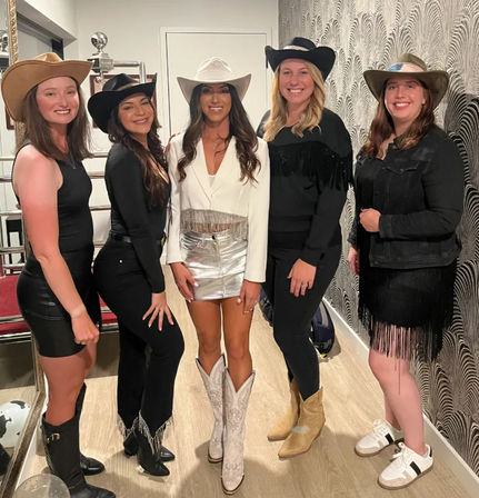 Five women in cowgirl-chic outfits wearing cowboy hats and boots, smiling and posing indoors in a modern hallway with patterned wallpaper and wood floor; center woman in a white blazer, silver mini skirt and white cowboy boots.