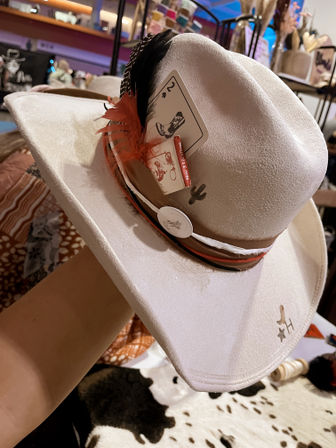 White felt cowboy hat with red and black feathers and a 2 of spades card tucked in the band — western rodeo-style hat with decorative band and cactus pin