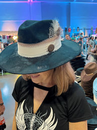 Woman trying on a stylish black wide-brim fedora with a cream fabric band, button accent and spotted feather at a busy indoor market stall