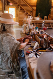 Person in a wide-brim hat decorating a felt hat at a wooden craft table in a rustic barn-style workshop, surrounded by ribbons, scissors, feathers and dried flower arrangements.