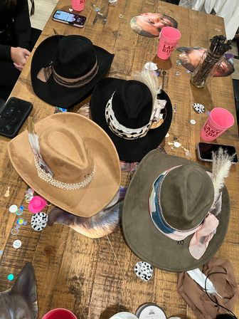 Overhead shot of a rustic wooden table with five decorated cowboy hats (black, tan, olive), feathers and patterned bands, pink party cups, confetti, and phones scattered around.