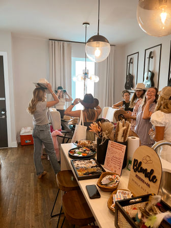 Friends trying on cowboy and western hats at a pop-up hat bar in a bright modern kitchen, guitars on the wall, pendant lights overhead, and an island covered with hat accessories and dried florals.