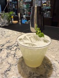 Pale green iced cocktail with mint garnish in a clear cup on a marble bar countertop, stainless cocktail shaker and liquor bottles blurred on the bar in the background.