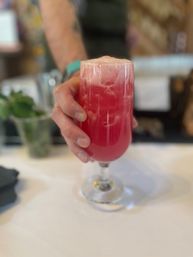 Hand holding a tall stemmed glass with a bright pink frothy cocktail on a white bar counter — fruity cocktail at a bar.