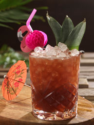 Tropical tiki cocktail in a faceted glass with crushed ice, pineapple-leaf garnish, pink flamingo straw and orange paper umbrella on a wooden table