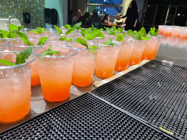 Rows of vibrant orange-pink iced cocktails in clear plastic cups, each topped with a fresh mint sprig, lined up on a bar counter at an indoor event