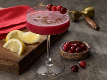 Bright cranberry cocktail in a coupe glass garnished with a cranberry skewer, lemon wedges on a wooden board and a jigger on a rustic tabletop.
