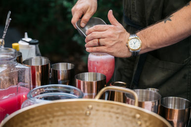Hands of a bartender pouring a frothy pink cocktail into a glass at an outdoor bar station, surrounded by copper shakers and pitchers