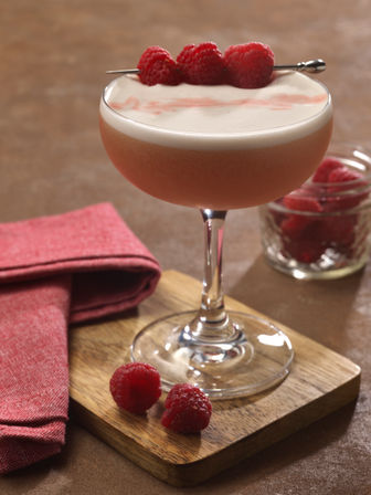 Frothy raspberry cocktail in a coupe glass, garnished with three raspberries on a skewer, placed on a wooden board with a red napkin and jar of raspberries