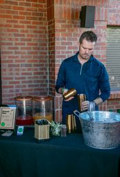 Person mixing mocktails at an outdoor brick-patio drink station — juice dispensers, copper shakers, a large galvanized ice bucket, garnish jars and a visible mocktail menu.