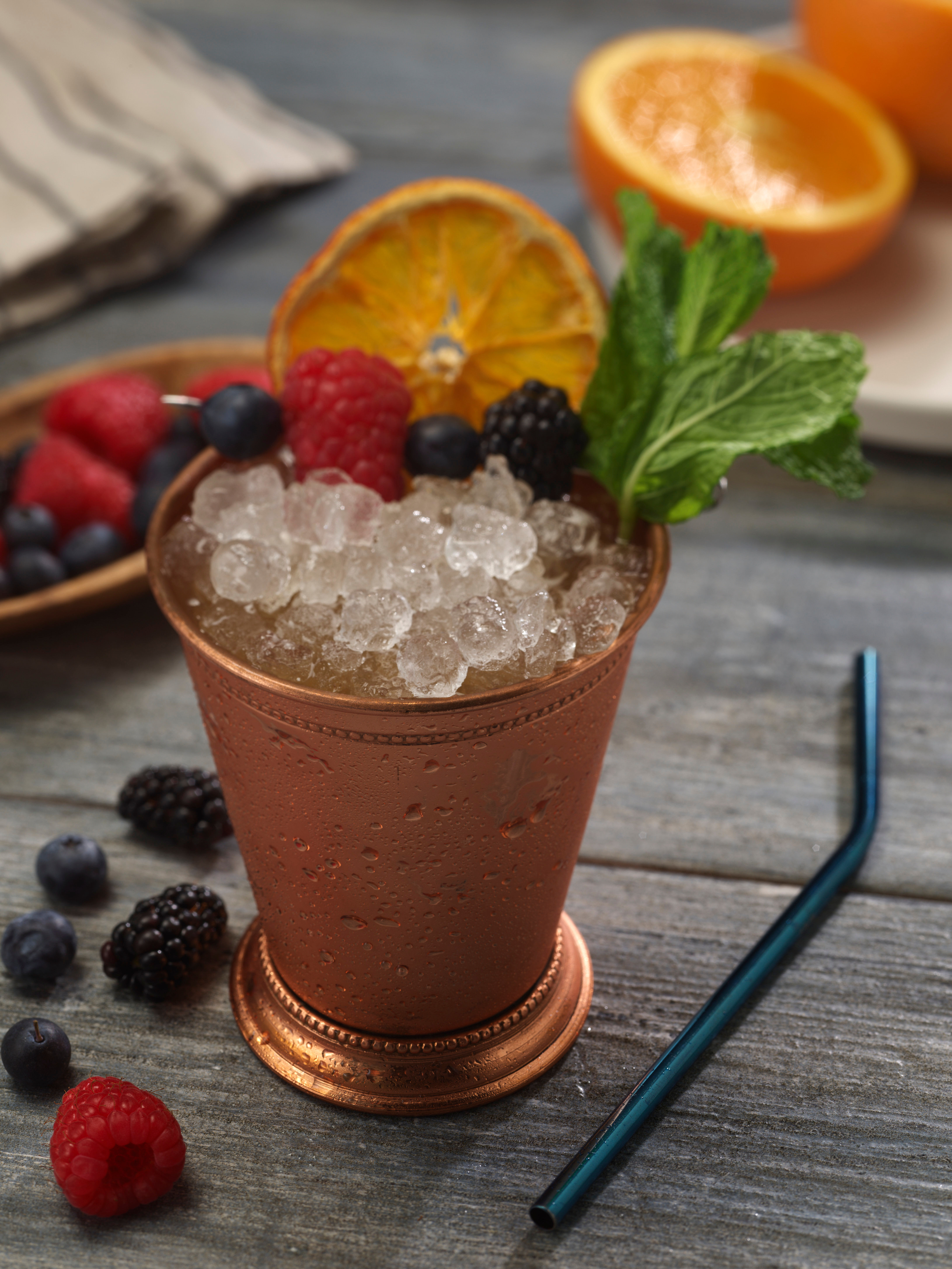 Frosty cocktail in a copper julep cup topped with crushed ice, fresh mint sprig, dried orange slice and mixed berries on a rustic wooden table