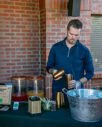Person preparing drinks at an outdoor brick-patio mocktail station with glass beverage dispensers, copper shakers, fresh herbs and a large metal ice bucket.