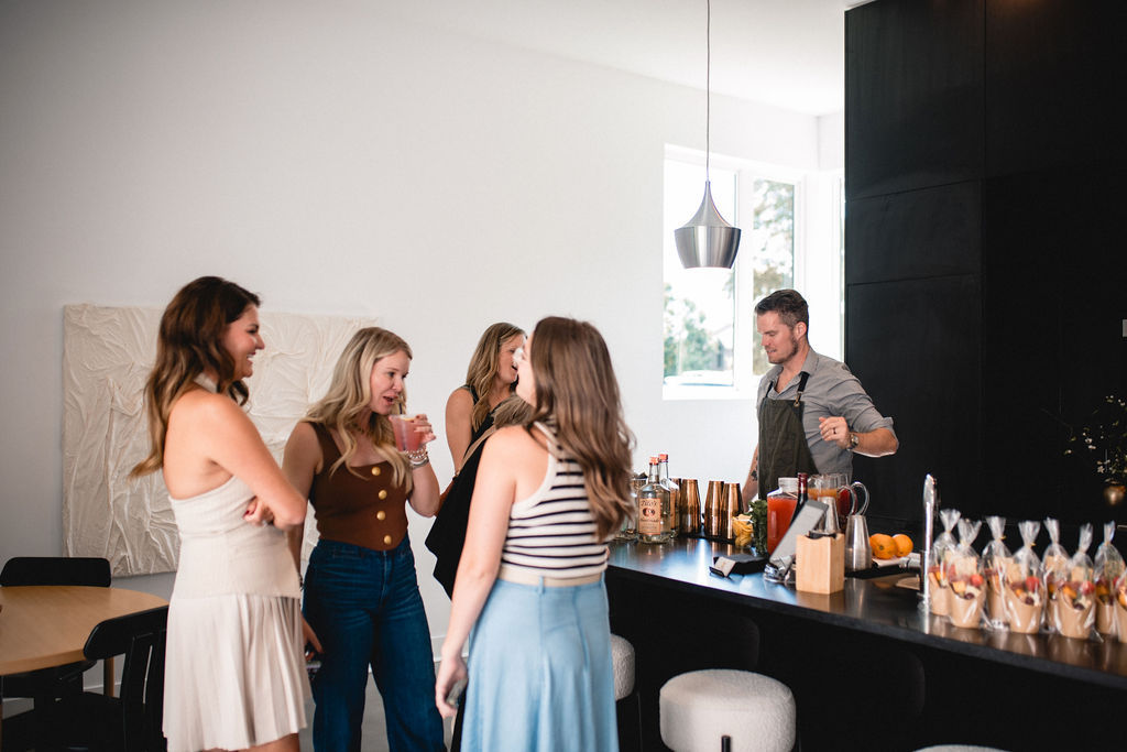 Four women chatting at a modern kitchen bar as a bartender mixes cocktails and arranges fruit cups for a casual home gathering.
