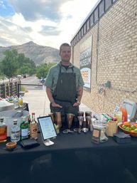 Outdoor mobile bar at a museum event in a mountain town — bartender in a green shirt and dark apron stands behind a table with cocktail shakers, liquor bottles, mixers, fresh citrus and a tip jar, brick building and foothills under a cloudy sky in the background.