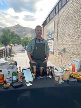 Outdoor mobile bar at a museum event in a mountain town — bartender in a green shirt and dark apron stands behind a table with cocktail shakers, liquor bottles, mixers, fresh citrus and a tip jar, brick building and foothills under a cloudy sky in the background.
