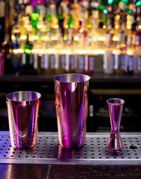 Iridescent metal cocktail shakers and a jigger on a perforated bar rail with colorful backlit liquor bottles in the background