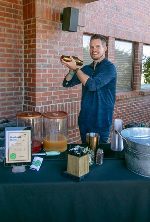 Smiling bartender shaking a copper cocktail shaker at an outdoor red brick patio drink station with beverage dispensers, plastic cups and a mocktail menu on the table.