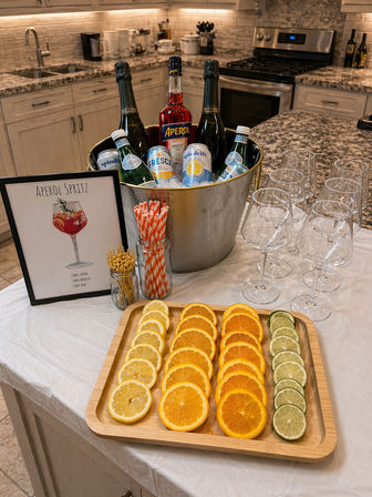 Aperol Spritz drink station on a kitchen island: ice-filled metal bucket with Aperol, sparkling bottles and mixers, lined wine glasses, striped paper straws, and a wooden tray of lemon, orange and lime slices for garnishes.