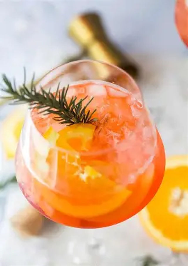 Top-down view of a zesty orange cocktail with crushed ice, orange slices and a rosemary sprig garnish in a stemmed glass on a marble surface