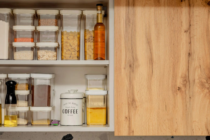 Organized kitchen pantry shelf with clear airtight containers of pasta, grains and lentils, a tall oil bottle and a white coffee canister inside a light wood cabinet.