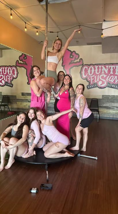 Cheerful group of women in pink and athletic wear posing on and around a pole in a bright dance-fitness studio.